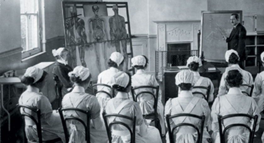 B+W photo of a nursing class being taught, there is a life size muscular-skeletal diagram 