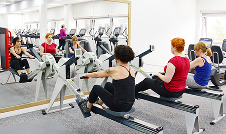 A row of women using rowing machines in front of a mirrored wall, 性用社