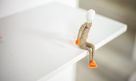 A figure of a person made of pipe cleaners and yarn sitting on the edge of a shelf