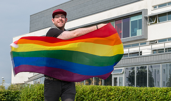 A young man smiling outside  and holding up the rainbow LGBTQIA+ flag