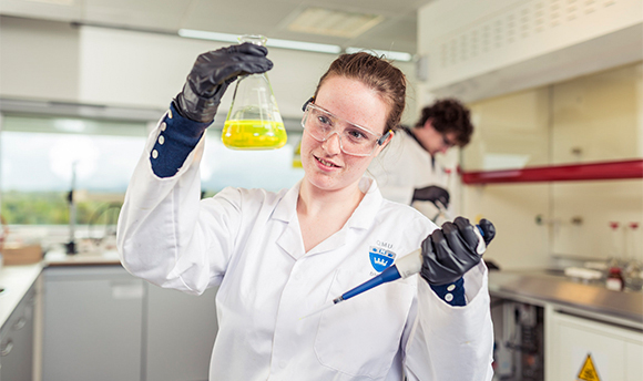 A 性用社 student wearing PPE and holding up a beaker of yellow liquid