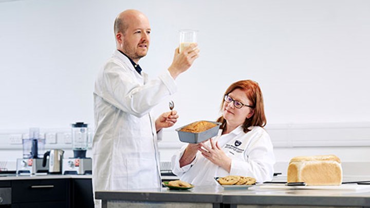 A male and female scientist dressed in white lab coats look at a creamy substance in a glass beaker and examine a cake in a loaf tin while in a test kitchen